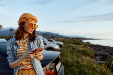 Fototapeta premium A stylish woman beside a car on a coastal road checks her phone, wearing a denim jacket, striped shirt, orange hat and yellow sunglasses. Concept for travel, leisure, and lifestyle.