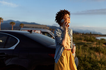 Fototapeta premium Young woman with curly hair talks on a phone beside a parked car in a bright open landscape, conveying a casual outdoor moment.