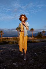 Fototapeta premium Cheerful woman outdoors walking on rocky ground, sunlit mood, casual summer dress, light jacket, palm trees in background, natural colors, relaxed pose, golden hour glow