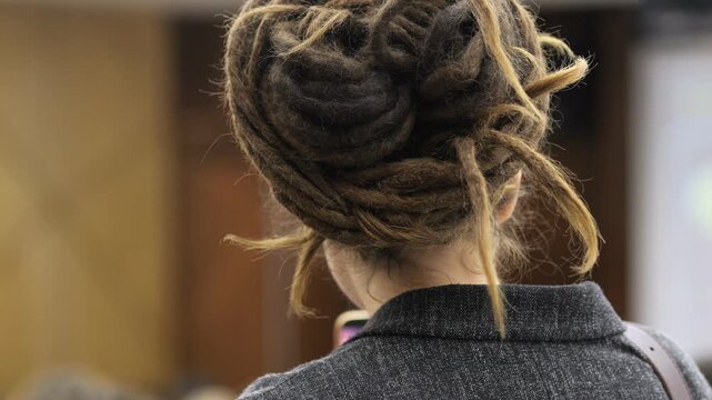 Back view of a person with styled dreadlocks, wearing a gray jacket. Hair texture shows natural curls and varied tones. Person appears calm and focused, possibly attending an event or meeting