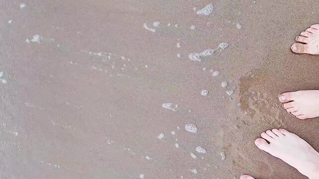Three pairs of bare feet standing on wet rocky shore as white sea foam waves wash in, top-down POV shot, Mallorca beach, family summer moment