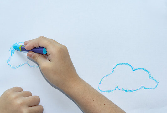 Child's hands drawing blue cloud outlines with oil pastel crayon on white paper, creative art activity, childhood education and learning.