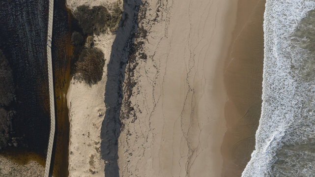 Aerial view of the Atlantic Ocean coastline featuring a sandy beach, rhythmic waves, and a wooden boardwalk path running alongside a dark water lagoon. Portugal.