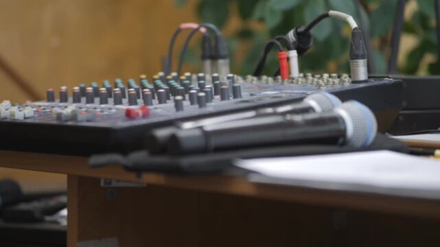 Close-up of audio mixing console with microphones on wooden table. Equipment ready for live sound event. Blurred greenery in background adds natural ambiance. Ideal for music, podcast