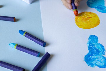 Child's hand coloring a yellow circle with an orange crayon on white paper, scattered blue crayons on light blue table, top view, creativity concept. © Irina