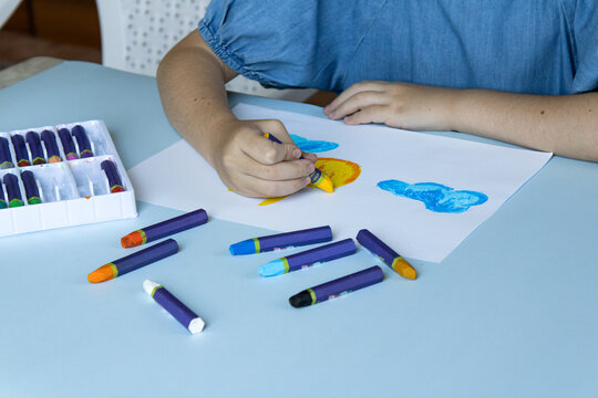 Child coloring yellow sun with oil pastel, white organizer box with sorted crayons on left, purple crayons with colored tips on light blue table, cloud drawing on white paper.