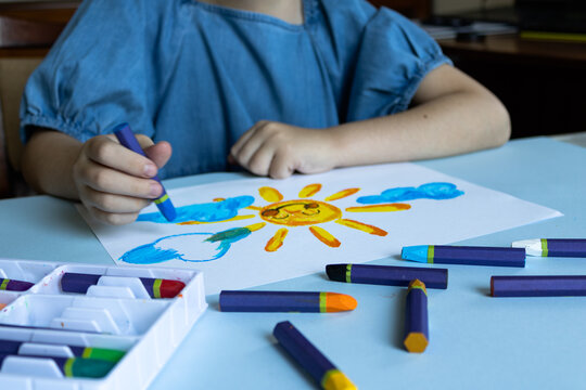 Child in blue shirt creating sun artwork with blue oil pastel, white paper with yellow sun and clouds, organized supplies in white box, scattered crayons on table.