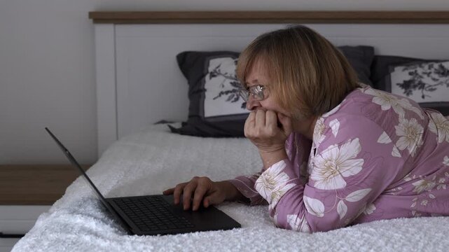 Senior woman lying on bed and using a laptop at home. Relaxed bedroom scene of an older woman browsing the internet, communicating online or enjoying digital leisure.