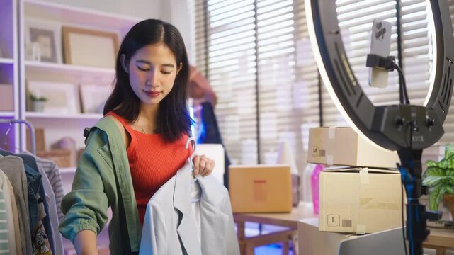 Asian woman live streaming fashion clothing sales with smartphone and ring light, modern retail e-commerce entrepreneur, social commerce influencer and generation z digital side hustle concept