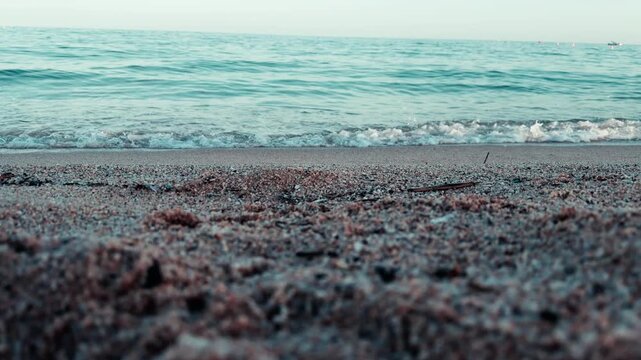 V&iacute;deo de una playa grabada a ras de suelo, con arena y peque&ntilde;os guijarros en primer plano y olas suaves llegando a la orilla. La escena muestra un paisaje costero tranquilo, con horizonte marino y luz