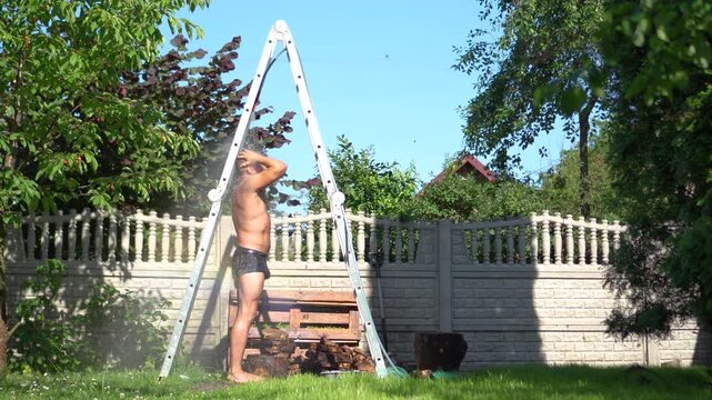 Wide commercial shot of an athletic man cooling down under a creative homemade DIY shower made from a garden hose and a stepladder in his backyard during the extreme European heatwave.