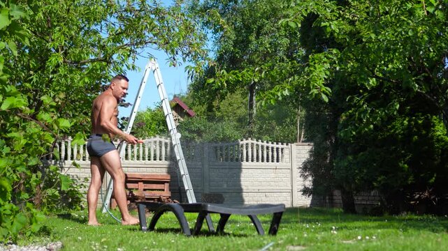 Wide commercial shot from the back of an athletic man in swimmer briefs using a garden hose to water the green lawn in his backyard during the extreme European heatwave.