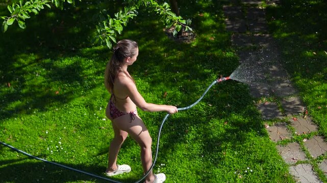 High angle commercial shot from a second floor balcony of a woman in a bikini using a garden hose to water the green lawn in her backyard during the extreme European heatwave.