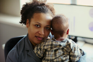 Fototapeta premium Portrait of African American young mother holding baby in office, balancing caregiving with work responsibilities. Ideal for working parent, maternity policy, hybrid work marketing