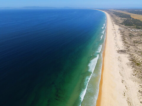 Aerial view of Praia da Comporta with turquoise waters, golden sand dunes, and gentle waves along the Atlantic coastline in Comporta, Setubal, Portugal.