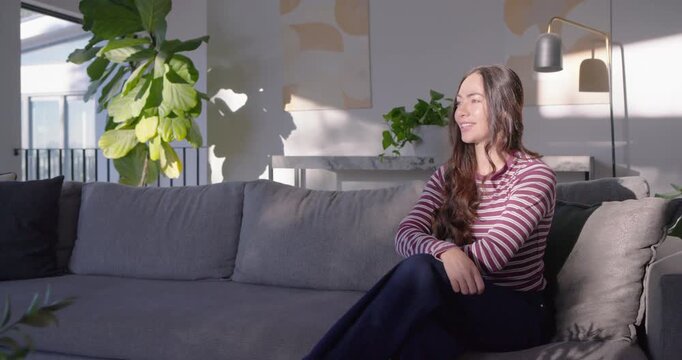 Adult woman sitting on grey sofa at home turning toward camera, reacting to off-camera cue