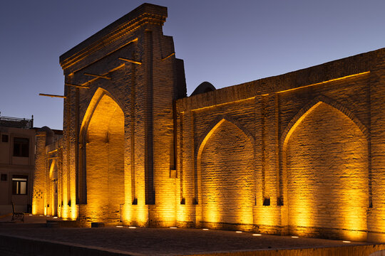 Historic brick facade with pointed arches illuminated at twilight in Ichan Kala old town of Khiva, Uzbekistan. Dramatic warm uplighting highlights the medieval Islamic architecture against a deep.