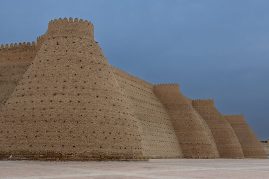 Ark Fortress walls in Bukhara with massive sloping buttresses and crenellated parapets under an overcast sky. This iconic Uzbekistan landmark image is ideal for travel publications and heritage.