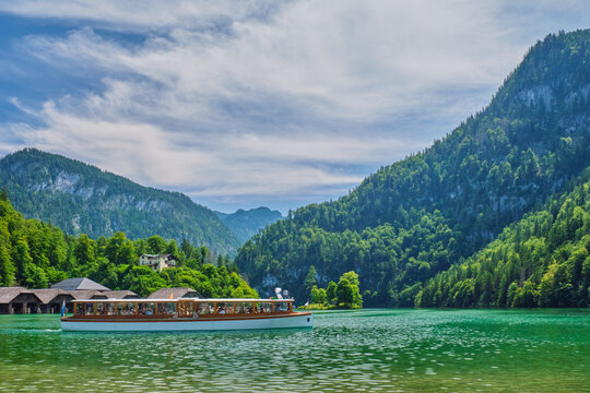 Side view of a white electric passenger boat cruising the emerald Konigsee lake under a dramatic sky in Berchtesgaden National Park, Bavaria, Germany.