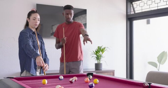 Diverse couple in denim, red tee, pointing and taking shot on pink-felt table cues balls scattering