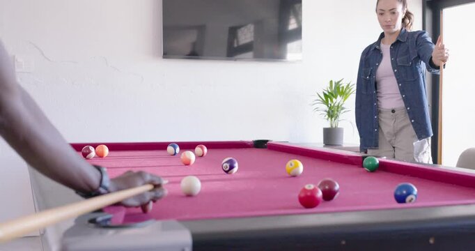 Diverse couple watching man stepping forward, striking cue ball on red table at home, celebrating
