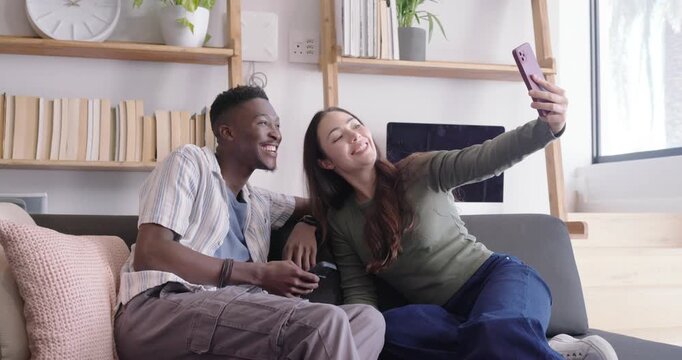 Diverse couple on gray sofa, woman raising pink phone, man with remote taking and reviewing selfies