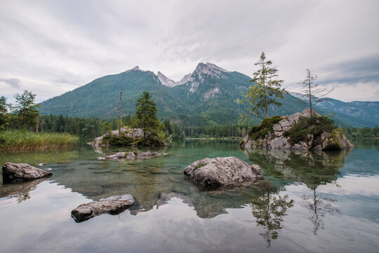 Wide angle view of the transparent water at Hintersee lake featuring rocky islands and the Hoher Goll mountain range in Germany.