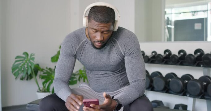 African American man in grey putting on headset tapping phone selecting music, nodding on gym bench