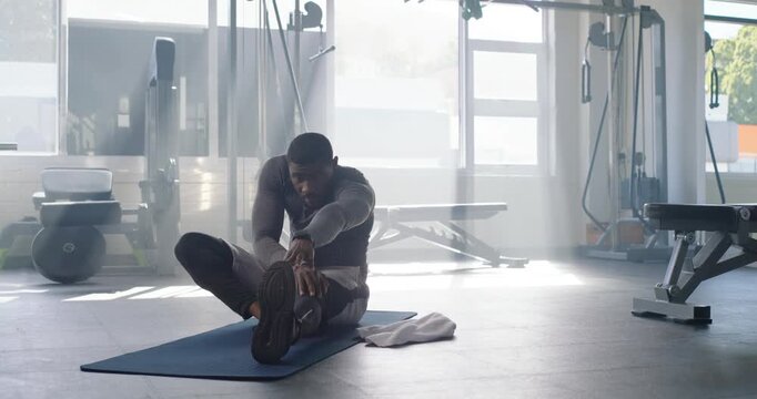 Mid-30s African-American man stretching hamstring on mat in gym, gray top, towel beside, warming up