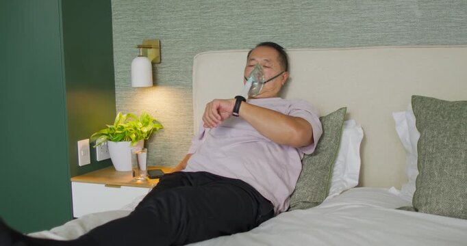 Senior Asian man checking smartwatch buzz, adjusting oxygen mask for breath, sitting upright on bed