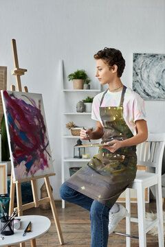 A young woman in an apron paints a colorful picture, fully immersed in her artistic hobby at home.