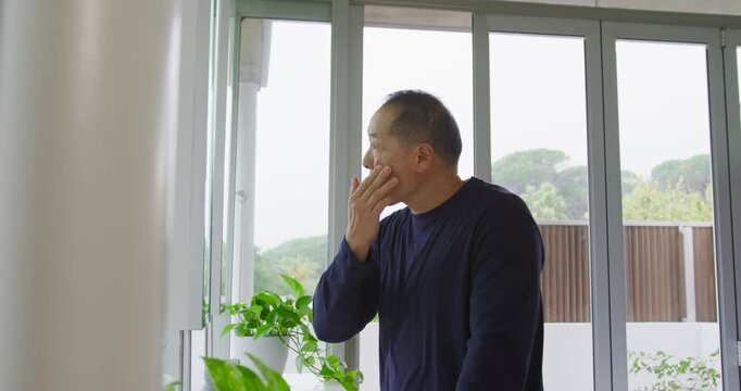 Senior Asian man by glass doors with plants seeing reflection, touching eye and inspecting face