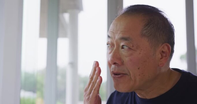 Senior Asian man noticing skin texture, touching cheek near window posts and greenery, copy space