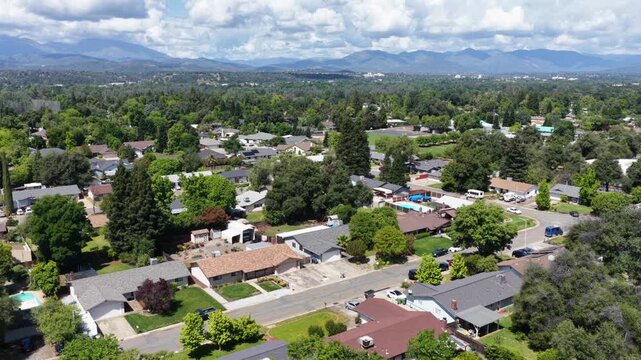 Redding suburban homes nestle among lush green trees, quiet streets winding through peaceful neighborhood beneath distant hazy California mountains on calm summer day. Aerial drone wide shot
