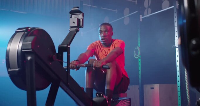 Adult man of African descent rowing on ergometer in gym following watch timer, wearing bright shirt