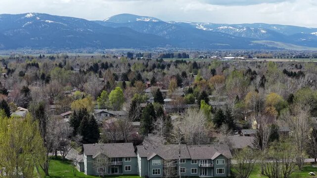 : Bozeman residential townhomes nestle among budding spring trees as snow-capped mountain peaks rise above quiet valley neighborhood and distant farmland. Montana, USA. Aerial drone wide shot