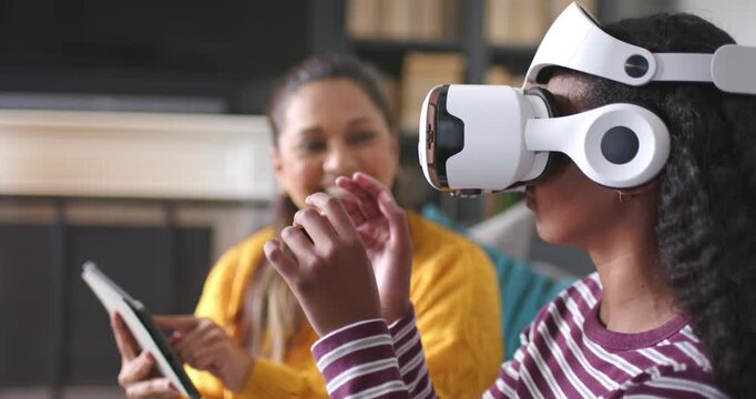 Diverse female mom and teen on couch, teen donning white VR headset, mom tapping tablet guiding
