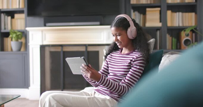 African American youth female sitting right on couch with pink headphones, tapping tablet, smiling