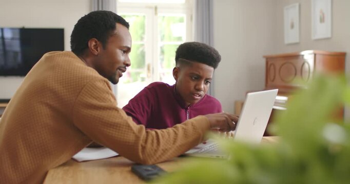 African American father and teen son guiding and learning at wooden table, adult pointing at laptop