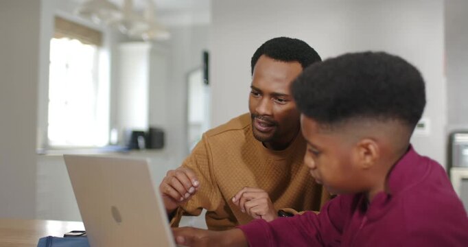 African American father guiding son at home, pointing green pencil while son typing laptop homework