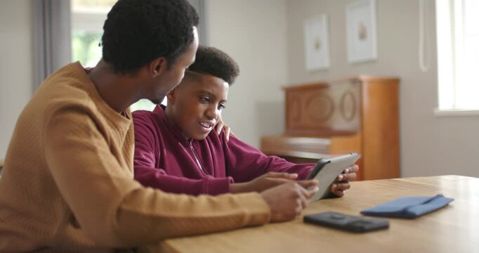 African American father guiding teen son in maroon hoodie at home, helping with tablet homework