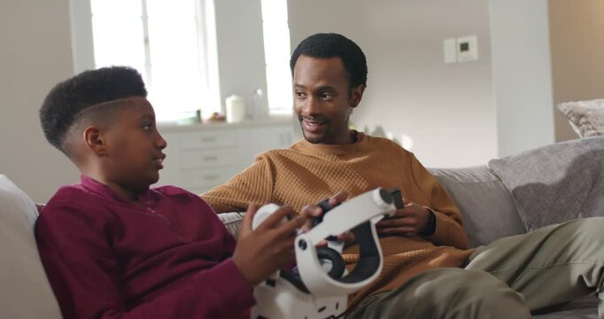 African American father guiding teen on couch, teen speaking while testing VR headset with tablet