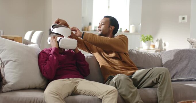 African American father and teen holding VR headset on couch, dad adjusting straps ensuring fit