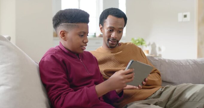 Father and teen son sitting on sofa, son showing tablet and tapping, father guiding son