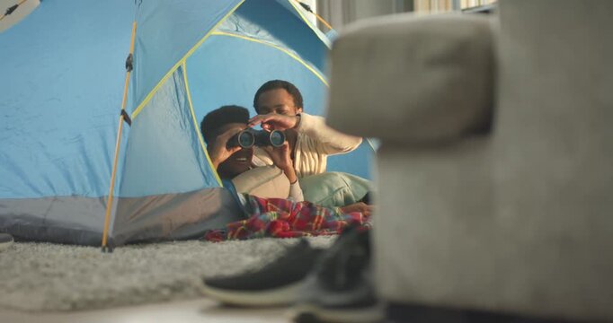 African American father and teenage son camping in blue tent at home, father steadying binoculars