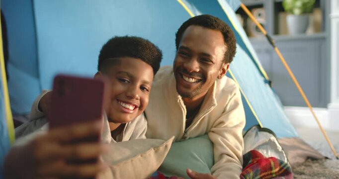 African American father and son posing in blue tent at home, child holding smartphone saving memory