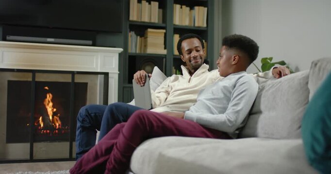 African American father and teen son reviewing schoolwork using tablet on couch by fireplace