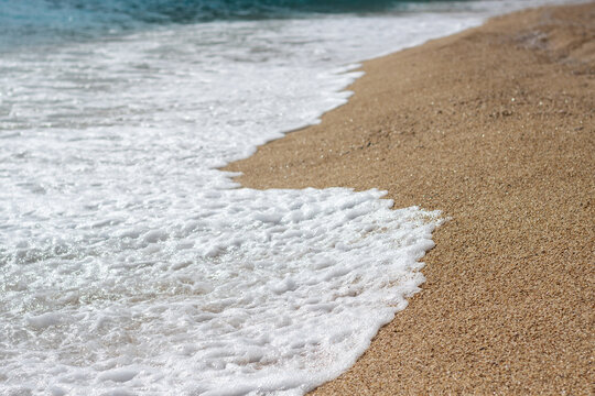Close-up top view of abstract white sea wave foam smoothly spreading across a golden pebble and sand beach at Kaputas, Turkey. Transparent water texture.