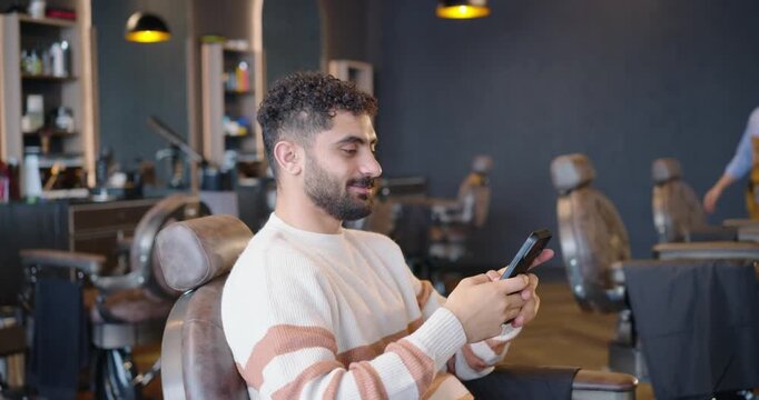 Two African men; client scrolling phone, barber walking in apron, greeting and styling hair in shop