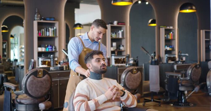 Male barber fitting neck strip and draping cape at barbershop while male client waiting for haircut